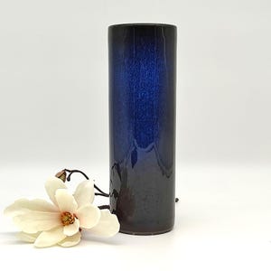 Könnte beinhalten: Eine hohe, zylindrische Vase mit Farbverlauf von tiefblau nach dunkelbraun. Eine weiße und gelbe Magnolienblüte liegt neben der Vase. Ein dekoratives Element für Blumen oder als Einzelstück.