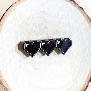 May include: A black pixelated heart pin set against a light wood slice. The pin features three black hearts with white accents, arranged horizontally. The wood slice is displayed on a black wire stand, creating a rustic yet modern aesthetic.