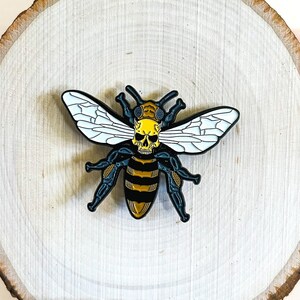 May include: A black and yellow enamel pin of a bee with a skull for a head. The bee is on a round wooden background.