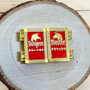 May include: Two gold-colored enamel pins with red backgrounds. The pins are hinged and feature a dragon design. The text on the pins reads "Dungeon Master" and "Dungeon".
