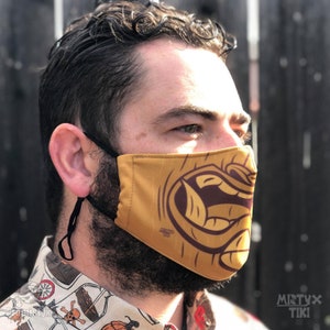 Pode incluir: Máscara facial amarela mostarda com um desenho tiki castanho. A máscara é usada por uma pessoa e tem presilhas pretas para as orelhas. A marca "Mistyx Tiki" é visível no canto inferior direito da imagem.