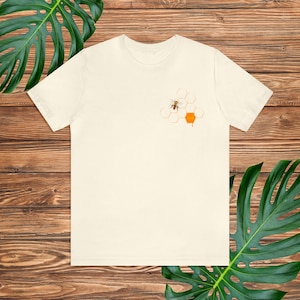 Könnte beinhalten: Cremefarbenes T-Shirt mit einer kleinen Grafik einer Biene und eines Bienenwaben-Designs. Die Biene ist schwarz und gelb, und die Bienenwabe ist orange. Das Shirt besteht aus einem weichen Material und hat einen klassischen Rundhalsausschnitt.