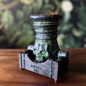 Puede incluir: Un tap&oacute;n de botella decorativo de aspecto oscuro y envejecido, con un dise&ntilde;o de calavera. El tap&oacute;n es de color verde oscuro y negro con un acabado texturizado. La base tiene el texto "ARTYX TIKI" en verde.