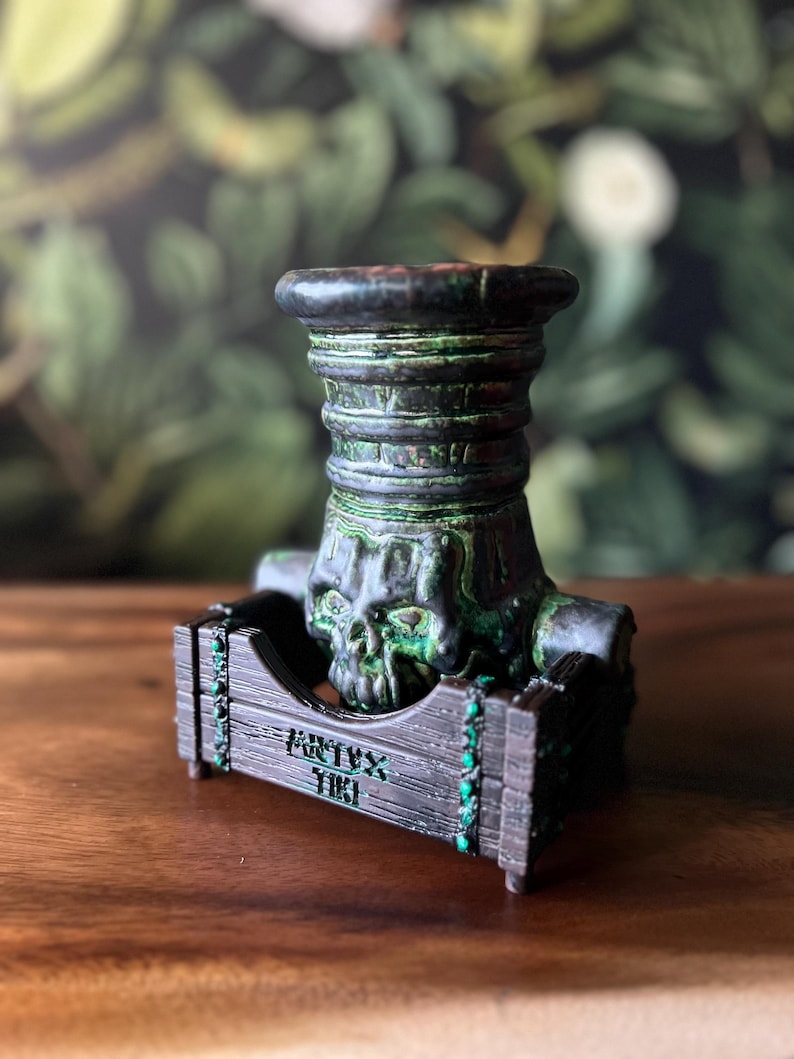 Puede incluir: Un tap&oacute;n de botella decorativo de aspecto oscuro y envejecido, con un dise&ntilde;o de calavera. El tap&oacute;n es de color verde oscuro y negro con un acabado texturizado. La base tiene el texto "ARTYX TIKI" en verde.