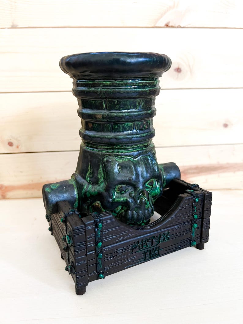 Puede incluir: Una pieza decorativa con tem&aacute;tica Tiki en verde oscuro y negro. El dise&ntilde;o presenta una calavera y una base de caja de madera con el texto "MATYX TIKI". La pieza tiene un aspecto texturizado y envejecido.