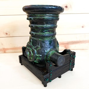 Puede incluir: Escultura decorativa de ca&ntilde;&oacute;n en verde oscuro y negro. Montada sobre una base de madera negra con detalles verdes. El ca&ntilde;&oacute;n presenta una superficie texturizada y un ca&ntilde;&oacute;n cil&iacute;ndrico.