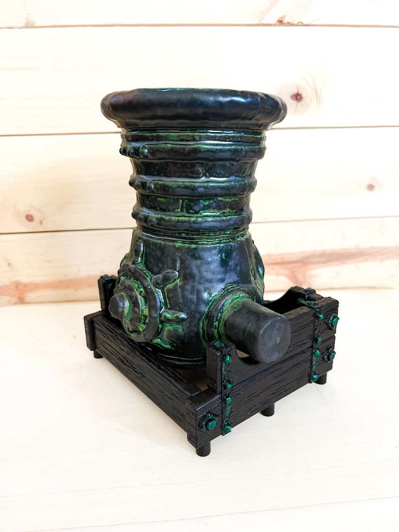 Puede incluir: Escultura decorativa de ca&ntilde;&oacute;n en verde oscuro y negro. Montada sobre una base de madera negra con detalles verdes. El ca&ntilde;&oacute;n presenta una superficie texturizada y un ca&ntilde;&oacute;n cil&iacute;ndrico.