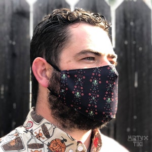 Könnte beinhalten: Schwarze Gesichtsmaske mit einem sich wiederholenden Muster aus grünen Tentakelfiguren, rosa Blumen und Totenköpfen. Die Maske hat schwarze elastische Ohrschlaufen. Der Hintergrund ist ein Holzzaun.