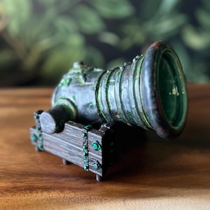 Puede incluir: Una escultura de ca&ntilde;&oacute;n en miniatura con un acabado met&aacute;lico envejecido. El ca&ntilde;&oacute;n presenta una cure&ntilde;a de madera marr&oacute;n oscuro con detalles verdes y un interior verde. El ca&ntilde;&oacute;n es de color verde oscuro y bronce.