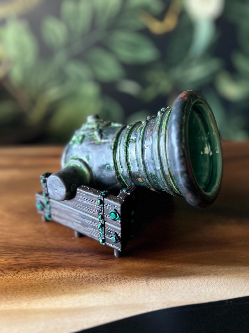 Puede incluir: Una escultura de ca&ntilde;&oacute;n en miniatura con un acabado met&aacute;lico envejecido. El ca&ntilde;&oacute;n presenta una cure&ntilde;a de madera marr&oacute;n oscuro con detalles verdes y un interior verde. El ca&ntilde;&oacute;n es de color verde oscuro y bronce.