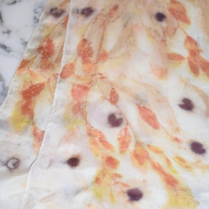 May include: A white silk scarf with a hand-painted design of leaves and branches in shades of orange, yellow, and brown. The scarf has a subtle texture and a few dark purple spots.