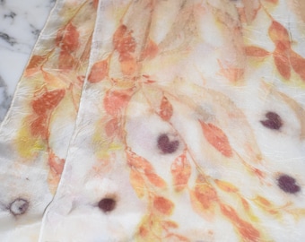 May include: A white silk scarf with a hand-painted design of leaves and branches in shades of orange, yellow, and brown. The scarf has a subtle texture and a few dark purple spots.