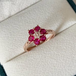 Op de afbeelding: Een roségouden ring met een bloemmotief. De bloem bestaat uit zeven ronde, dieprode edelstenen die een cluster van kleinere, heldere stenen omcirkelen. De ring wordt gepresenteerd in een witte sieradendoos met een turquoise interieur.