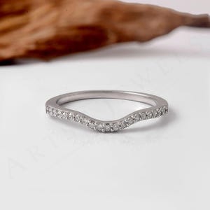 May include: A silver-toned curved band ring featuring a row of small, clear, round-cut stones set along the front arc. The metal has a polished finish, and the design follows a gentle wave shape, showcasing a series of sparkling accents.
