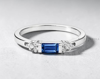 May include: A silver-toned ring featuring a central rectangular baguette-cut blue gemstone flanked by two clusters of three small round clear stones on each side. The band is smooth and polished, with the text Artsy Jewels visible on the right.