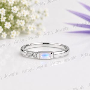 Op de afbeelding: Een zilverkleurige ring met een centrale rechthoekige maansteen in baguette-slijpvorm met een subtiele blauwe iriserende glans, aan één kant geflankeerd door drie kleine ronde heldere stenen gezet in de gepolijste metalen band.