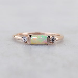 Puede incluir: Un delicado anillo de oro rosa con una piedra de ópalo rectangular y dos piedras redondas transparentes más pequeñas a cada lado.