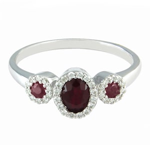 Op de afbeelding: Een zilveren ring met drie rode edelstenen. De middelste steen is ovaalvormig en omgeven door kleinere witte edelstenen. De twee kleinere stenen aan weerszijden zijn rond.