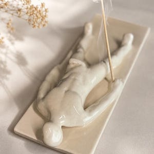 Könnte beinhalten: Ein cremefarbener Räucherstäbchenhalter aus Keramik mit einer skulptierten menschlichen Figur, die flach auf einer rechteckigen Basis liegt. Die Figur ist mit dem Gesicht nach unten positioniert und die Arme liegen seitlich. Ein Stäbchen steckt darin.