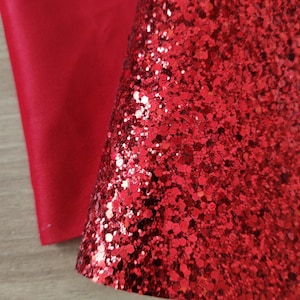 Peut inclure: Gros plan sur deux feuilles de matériau rouge. L'une est rouge uni mat, l'autre est recouverte de paillettes rouges scintillantes. La feuille de paillettes est pliée, révélant la texture et l'éclat des paillettes.