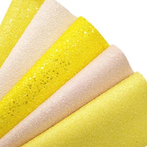 Op de afbeelding: Close-up van verschillende vellen getextureerd knutselpapier in tinten geel en gebroken wit. Het papier heeft een glitterende, sprankelende afwerking, met verschillende dichtheden van glitter. De vellen zijn uitgespreid en tonen de verschillende kleuren en texturen.