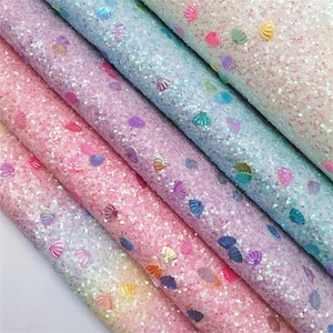 Pode incluir: Close-up de vários rolos de tecido com glitter em tons de rosa, roxo e azul. O tecido é coberto com pequenas lantejoulas brilhantes e enfeites em forma de concha. Ideal para artesanato e projetos decorativos.