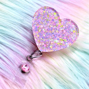 May include: A heart-shaped badge reel with a glittery, iridescent design in shades of pink, purple, and yellow. The badge reel has a clear plastic backing and a metal clip. The background is a soft, fluffy fabric in pastel colours.