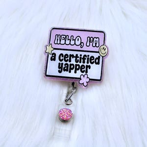 May include: A pink and white badge reel with the text "HELLO, I'M a certified yapper". The badge has a glittery border and is decorated with a star, a flower, and a smiley face. The reel has a pink, glittery button.