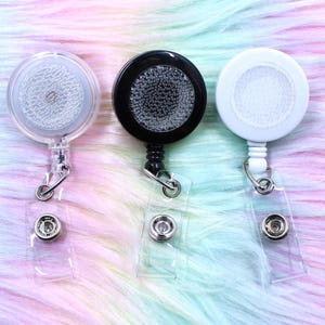 May include: Three retractable badge holders in clear, black, and white. Each has a round, textured top and a clear plastic strap with a snap closure. The background is a soft, multi-colored faux fur.
