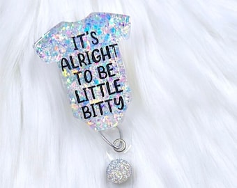 Könnte beinhalten: Ein glitzernder silberner Glitzer-Badge-Rollenhalter mit einem durchsichtigen Plastik-Badge-Halter. Der Anhänger ist in Form eines Stramplers mit dem Text "It's alright to be little bitty" gestaltet.