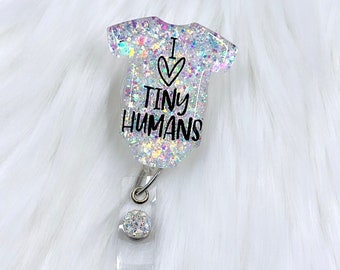 May include: A sparkly iridescent white glitter badge reel with a clear plastic baby onesie charm. The onesie has a pink heart with a white outline and the text "I ♥ TINY HUMANS".