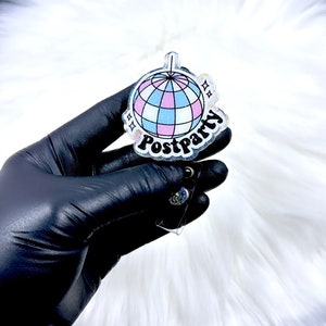 May include: A clear acrylic badge reel featuring a disco ball graphic with light blue, pink, and white panels. The word Postparty is printed in bold black lettering below the disco ball, accented by small star shapes and a subtle glitter finish.