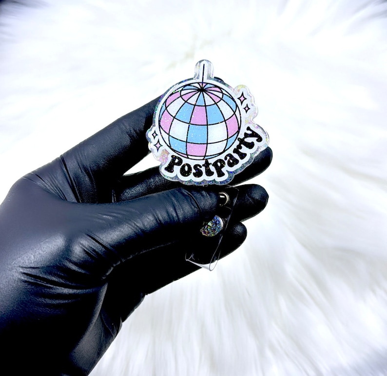 May include: A clear acrylic badge reel featuring a disco ball graphic with light blue, pink, and white panels. The word Postparty is printed in bold black lettering below the disco ball, accented by small star shapes and a subtle glitter finish.