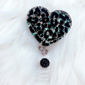Puede incluir: Un portagafete en forma de coraz&oacute;n negro y turquesa con un dise&ntilde;o de estampado de leopardo y un bot&oacute;n de pedrer&iacute;a negro.