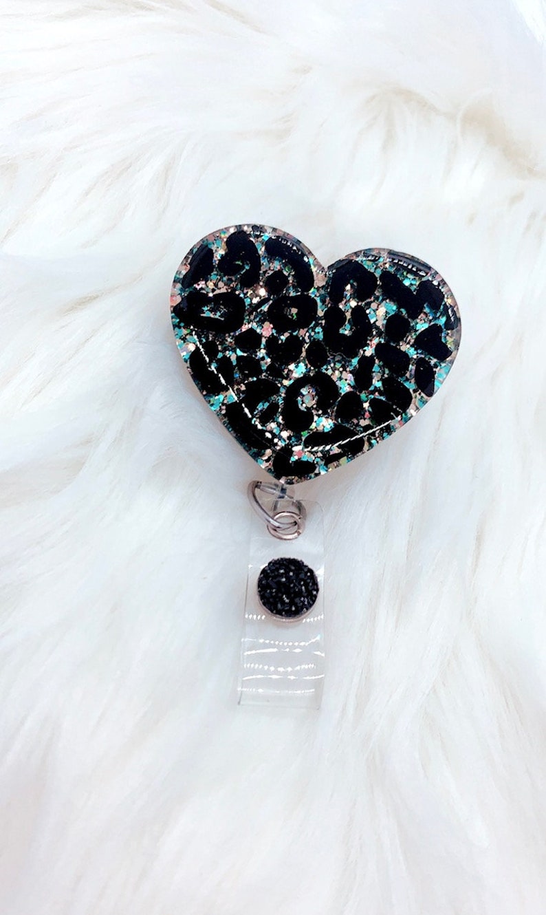 Puede incluir: Un portagafete en forma de coraz&oacute;n negro y turquesa con un dise&ntilde;o de estampado de leopardo y un bot&oacute;n de pedrer&iacute;a negro.