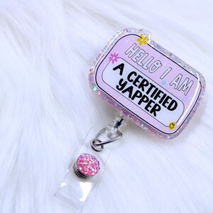 May include: Pink glitter badge holder with a clear retractable reel and a pink glitter button. The badge holder has the text "Hello I am a certified yapper" on it.