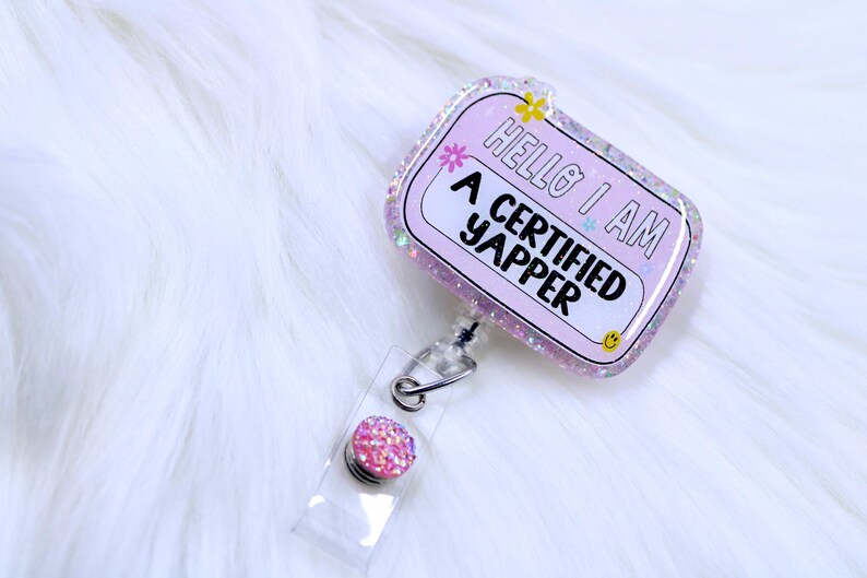 May include: Pink glitter badge holder with a clear retractable reel and a pink glitter button. The badge holder has the text "Hello I am a certified yapper" on it.
