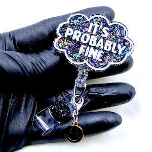 Pu&ograve; includere: Un portabadge a forma di nuvola con la scritta "IT'S PROBABLY FINE" in bianco. Il badge ha un design glitterato e colorato e un ciondolo nero e glitterato. Un piccolo ciondolo a forma di faccina sorridente dorata &egrave; appeso sotto.