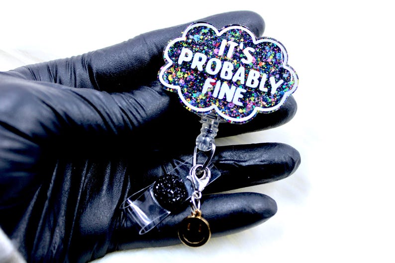 Pu&ograve; includere: Un portabadge a forma di nuvola con la scritta "IT'S PROBABLY FINE" in bianco. Il badge ha un design glitterato e colorato e un ciondolo nero e glitterato. Un piccolo ciondolo a forma di faccina sorridente dorata &egrave; appeso sotto.