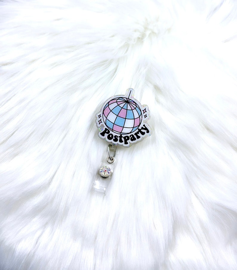 May include: A circular badge reel featuring a graphic of a disco ball with pink, blue, and white segments. The word Postparty is printed in black script lettering below the disco ball. The reel includes a clear plastic strap and a small rhinestone.