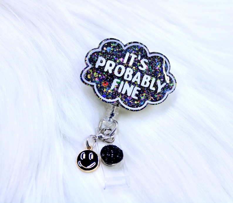 Pu&ograve; includere: Un portabadge con un design a forma di nuvola che recita "IT'S PROBABLY FINE" in lettere bianche. La nuvola &egrave; nera con glitter colorati. Un ciondolo a forma di faccina sorridente e una perla nera pendono dal portabadge.