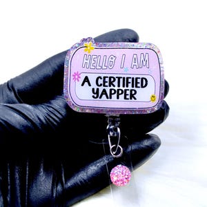 May include: Pink glitter badge holder with a clear plastic strap and a pink glitter charm. The badge holder has the text "Hello I am a certified yapper" printed on it.