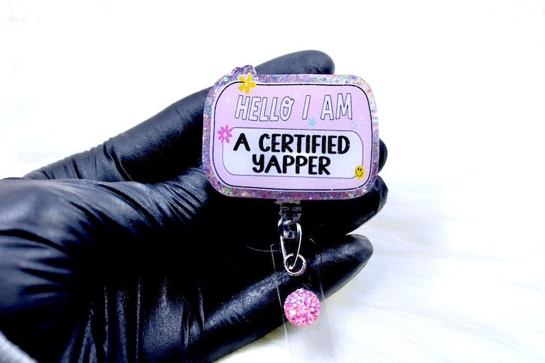 May include: Pink glitter badge holder with a clear plastic strap and a pink glitter charm. The badge holder has the text "Hello I am a certified yapper" printed on it.