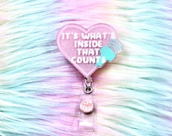 May include: A heart-shaped badge reel with a glittery pink finish. The text IT'S WHAT'S INSIDE THAT COUNTS is printed in white, accompanied by a small light blue ultrasound probe icon. The badge reel features a clear plastic strap and a metal clip.