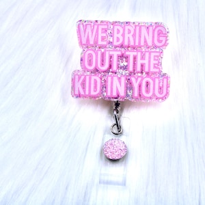 Op de afbeelding: Roze glitter badge reel met een witte achtergrond. De badge reel heeft een roze glitter charme met de tekst "We bring out the kid in you".