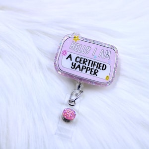 May include: Pink glitter badge holder with a clear retractable cord and a pink glitter charm. The badge holder has a pink background with white text that reads "Hello I am a certified yapper".