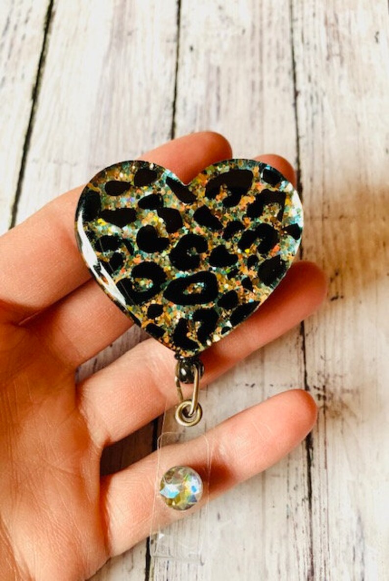 Puede incluir: Un portagafete en forma de coraz&oacute;n con estampado de leopardo, con un carrete de gafete retr&aacute;ctil transparente y un dije de cristal brillante.