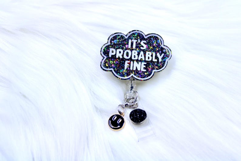 Pu&ograve; includere: Un portabadge retrattile con un design a forma di nuvola che recita "IT'S PROBABLY FINE" in testo bianco. La nuvola &egrave; nera con glitter colorati. Un ciondolo a forma di faccina sorridente e una sfera di glitter neri pendono dal portabadge.