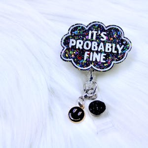 Pu&ograve; includere: Un porta badge con un design a forma di nuvola che recita "IT'S PROBABLY FINE" in lettere bianche. La nuvola &egrave; riempita di glitter colorati. Un ciondolo a forma di faccina sorridente dorata e una pallina di glitter neri pendono dal porta badge.
