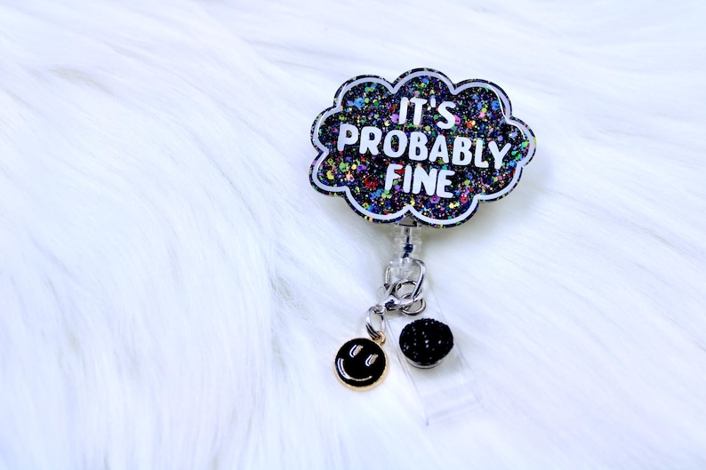 Pu&ograve; includere: Un porta badge con un design a forma di nuvola che recita "IT'S PROBABLY FINE" in lettere bianche. La nuvola &egrave; riempita di glitter colorati. Un ciondolo a forma di faccina sorridente dorata e una pallina di glitter neri pendono dal porta badge.